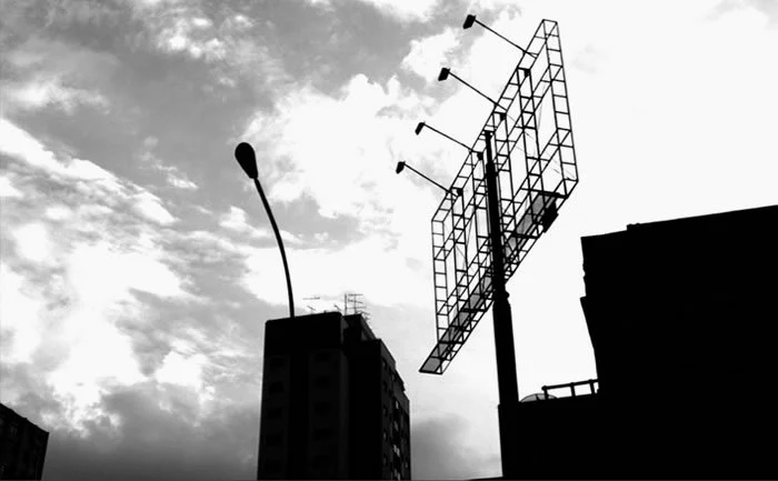 Black and white photo of an empty billboard frame against the sky, beside a street lamp and apartment blocks
